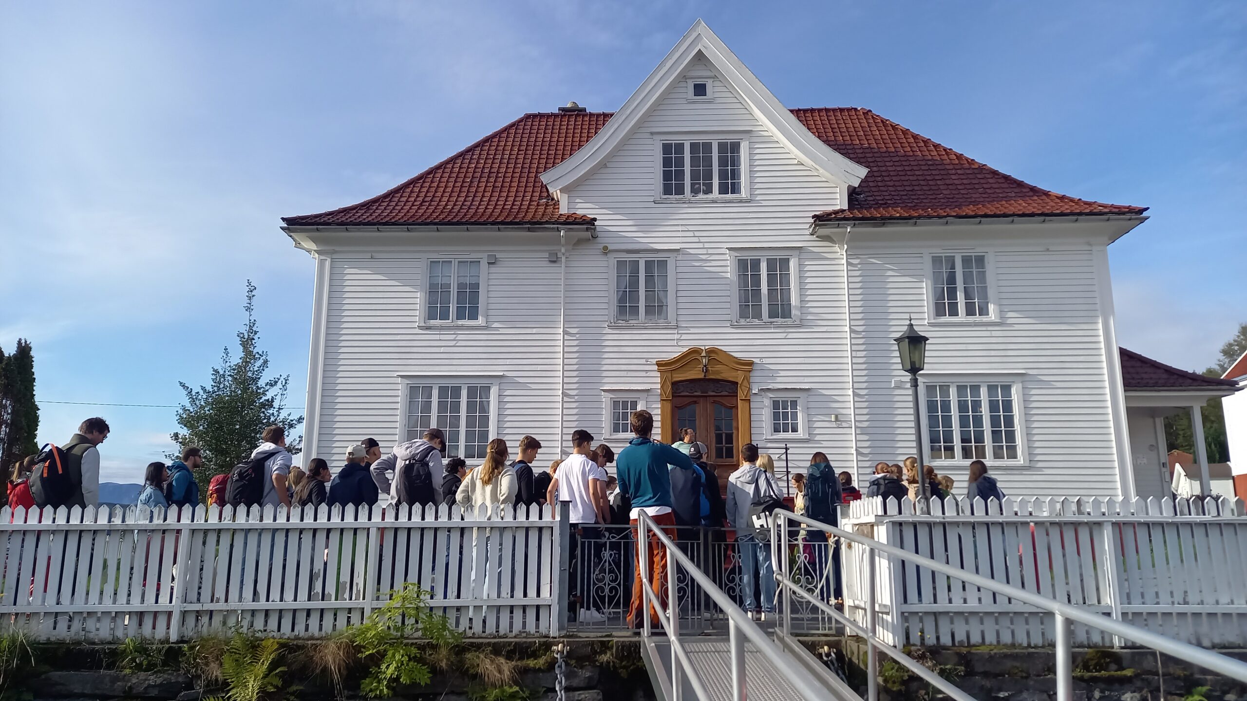 stort hvitt bygg med rødt tak. flere mennesker er plasser foran bygget